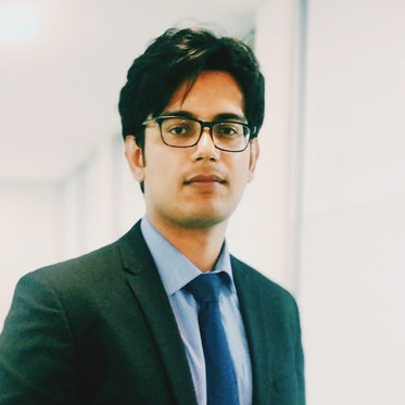 Person in a suit and tie, wearing glasses, standing indoors with a neutral expression.