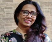 Person smiling, wearing glasses and a floral top, standing in front of a patterned wall.