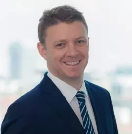 Person in a blue suit and striped tie smiling against a blurred cityscape background.