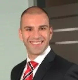 Person in a suit smiling, standing indoors with a blurred background.