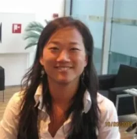 Person smiling indoors with a plant and chairs in the background.