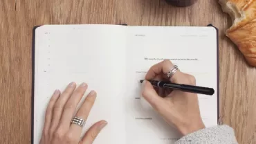 Person writing in a planner with a pen, near a croissant and a drink on a wooden table.