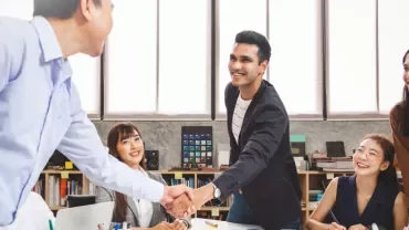 Group of people in an office, two shaking hands, others smiling at a desk.