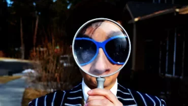 Person holds a magnifying glass in front of their face wearing sunglasses.