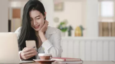 Person smiling at a phone in a cozy workspace with a laptop and coffee.