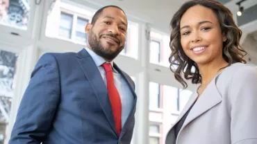 Two people smiling in a bright office with large windows.
