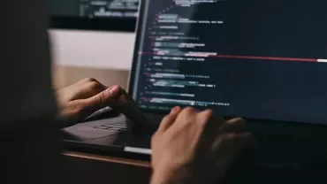 Person typing on a laptop with code on the screen in a dimly lit room.