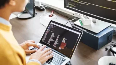Person coding on a laptop with screen monitors nearby.
