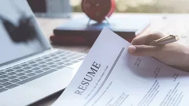 Person holding a resume next to a laptop on a desk.