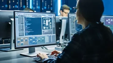 Person working at a computer with multiple monitors displaying code in a dimly lit office.