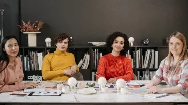 Four people sitting at a table in an office setting, surrounded by books and creative materials.