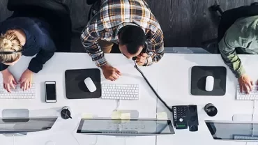 Overhead view of three people working at office desks with computers and phones.