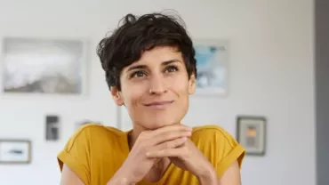 Person in a yellow shirt smiling indoors, resting chin on hands.