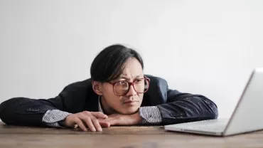 Person with glasses looking intently at a laptop on a wooden table.