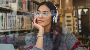 Person wearing headphones, seated by a library window with books in the background.