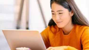 Person in orange sweater smiling while using a laptop.