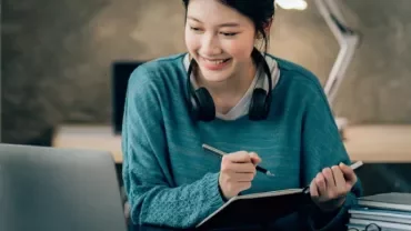 Person smiling at a laptop while taking notes at a desk.