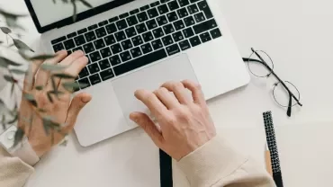 Person using a laptop with a notebook and glasses nearby on a desk.
