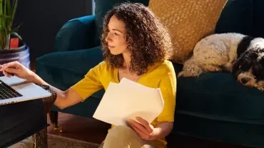 Person works on laptop with papers, dog rests on couch.