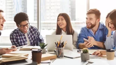 Group of people laughing and working together at a table with laptops and coffee.