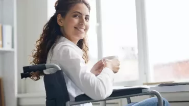 Person sitting in a wheelchair, smiling and holding a cup, near a laptop by a window.