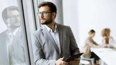 Person in suit holding tablet, standing by a window.