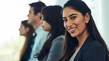 A woman in business attire smiles at the camera, sitting in a row with three other people, all facing forward in a professional setting.