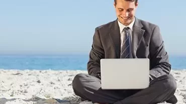 Person in a suit working on a laptop while sitting on a beach.