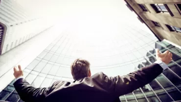 Person in a suit with arms raised towards a skyscraper.