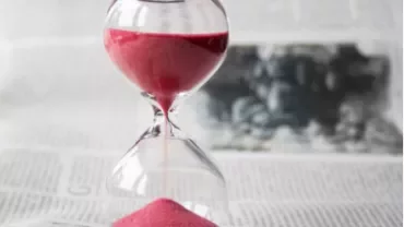 Red sand flows through an hourglass on a newspaper background.