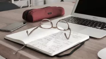 Notebook with glasses, pencil case, and a laptop on a wooden desk.