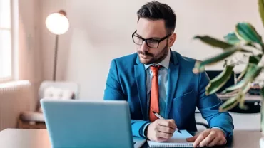 Person in blue suit works at laptop, jotting notes in a notebook.