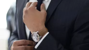 Person adjusting a tie while wearing a navy suit and a watch with a brown leather strap.