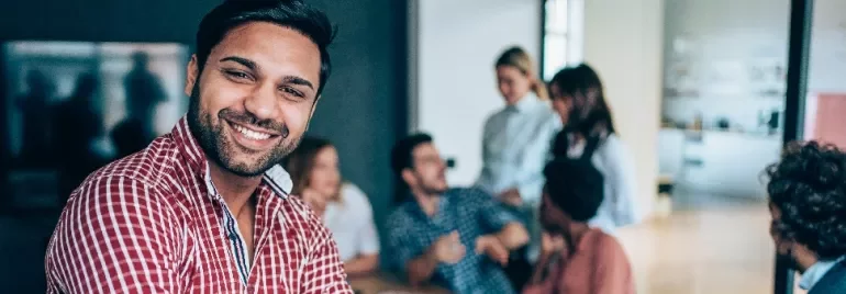 Smiling person in a checkered shirt stands in front of others in a modern office setting.