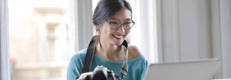 Cheerful woman with camera using laptop. Andrea Piacquadio via Pexels