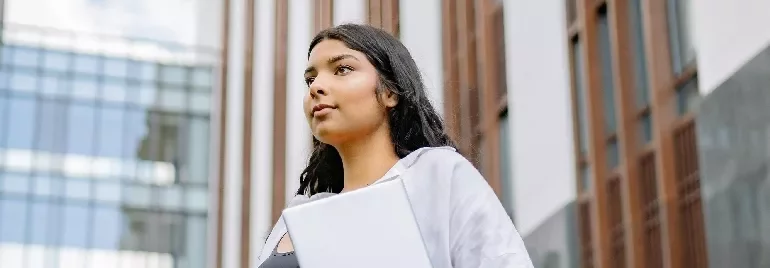 Person holding a laptop, standing in front of modern building.