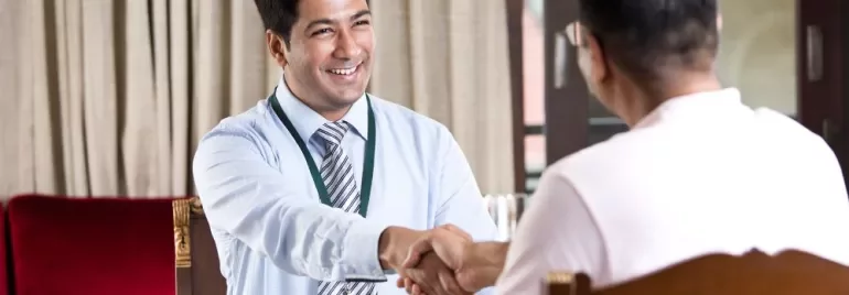 Two men sit across from each other at a desk, smiling and shaking hands in a well-lit office setting.