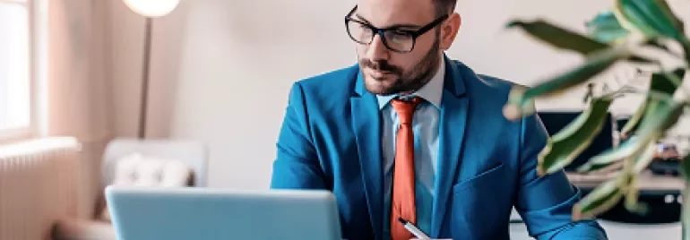 Person in blue suit works at laptop, jotting notes in a notebook.