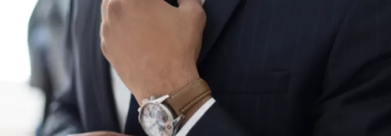 Person adjusting a tie while wearing a navy suit and a watch with a brown leather strap.