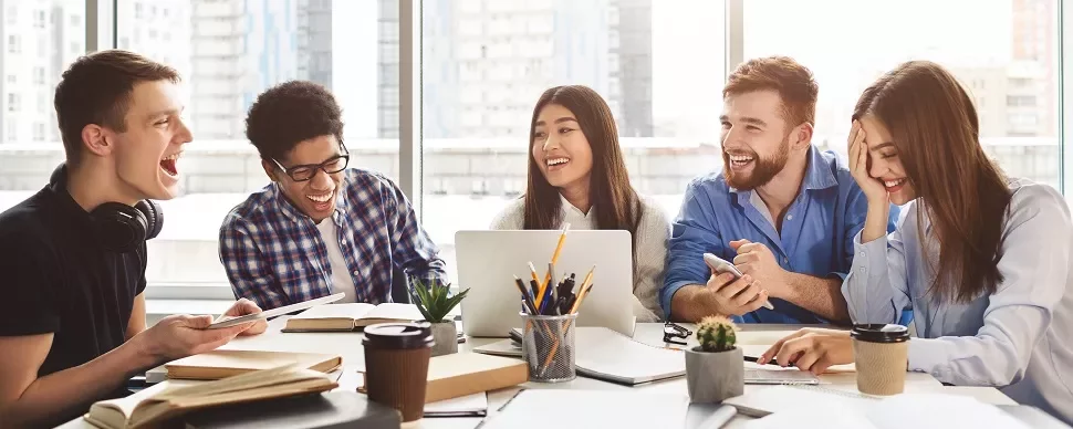 Group of people laughing and working together at a table with laptops and coffee.