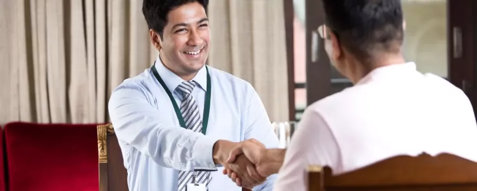 Two men sit across from each other at a desk, smiling and shaking hands in a well-lit office setting.