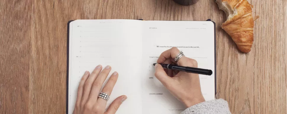 Person writing in a planner with a pen, near a croissant and a drink on a wooden table.