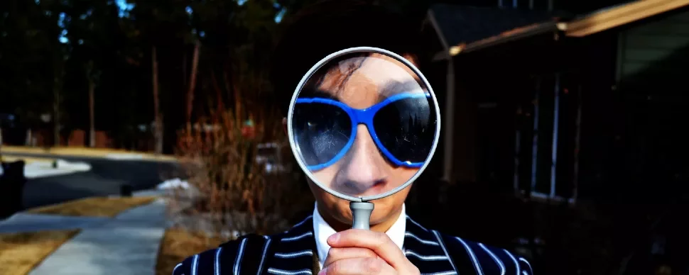 Person holds a magnifying glass in front of their face wearing sunglasses.