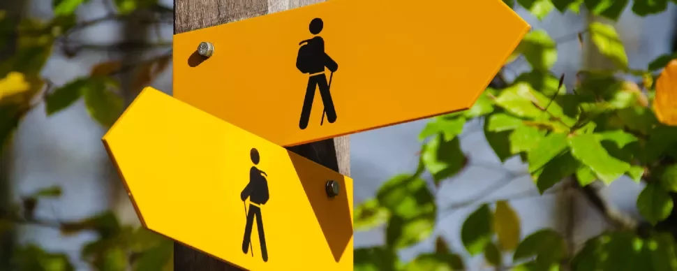 Two yellow hiking trail signs with arrows and backpack icons on a wooden post.