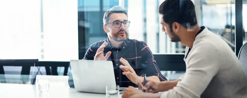 Two people having a discussion at a desk with a laptop and notepad.