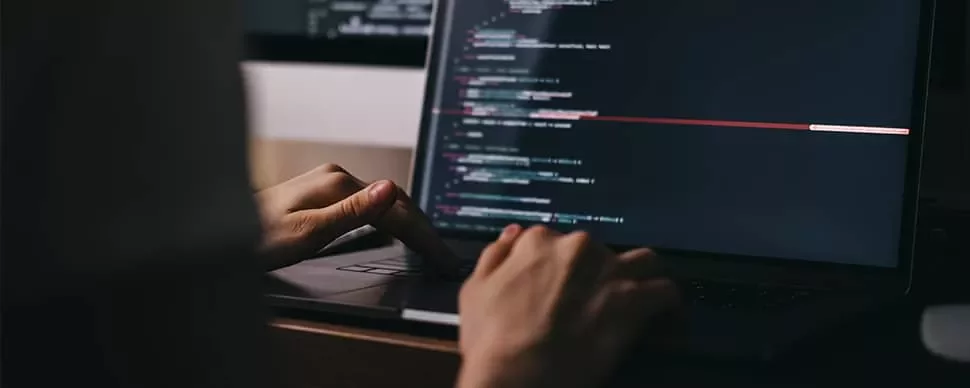 Person typing on a laptop with code on the screen in a dimly lit room.