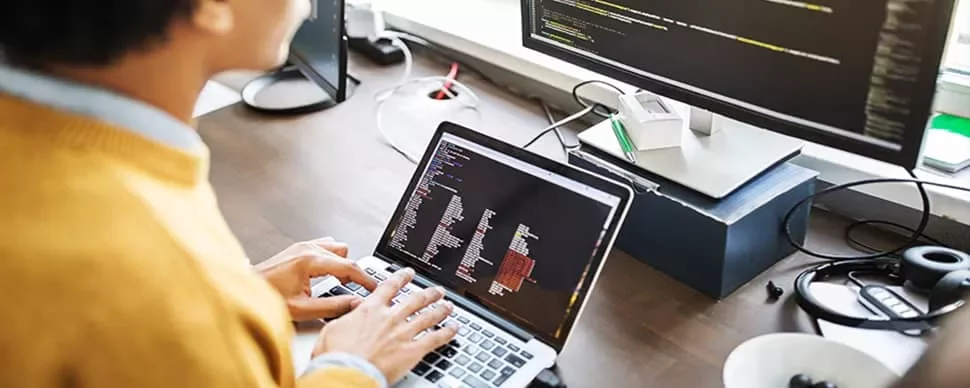 Person coding on a laptop with screen monitors nearby.