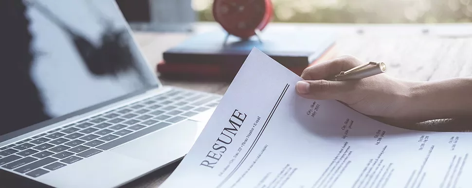 Person holding a resume next to a laptop on a desk.