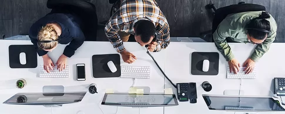 Overhead view of three people working at office desks with computers and phones.