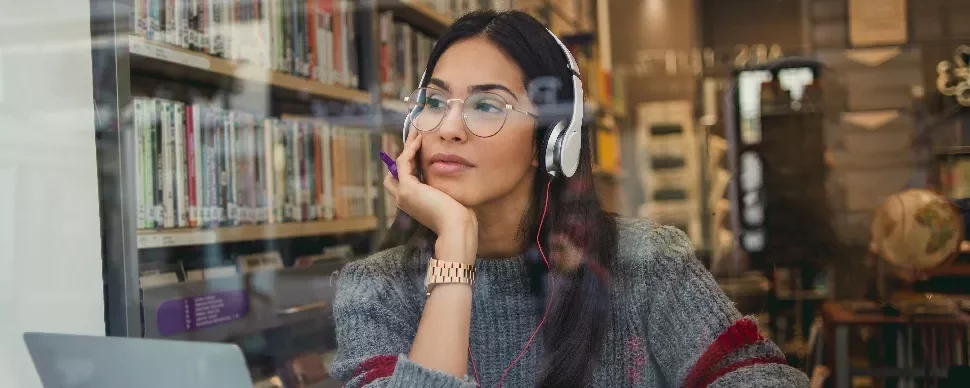 Person wearing headphones, seated by a library window with books in the background.